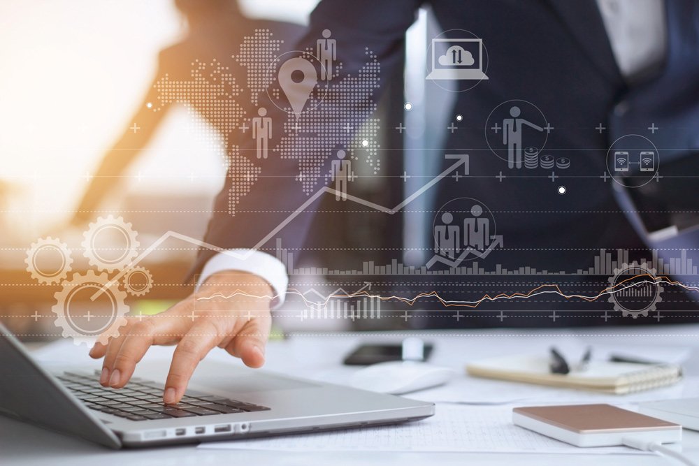 13 Infographic of world map, people icons and arrows and data transparently imposed on a background of a person in formal wear with arm touching a laptop's keyboard on a table with other items